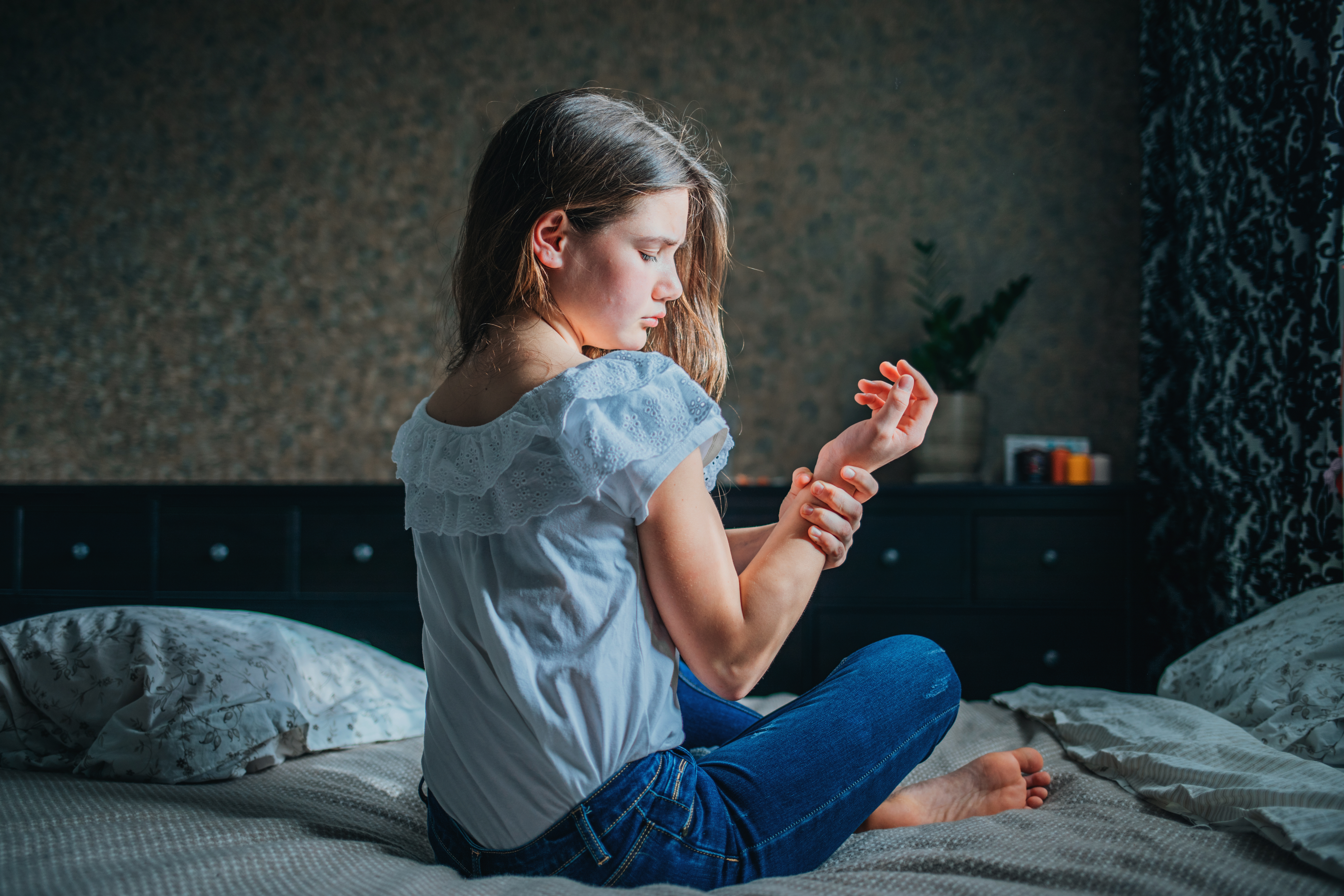 young dark-haired girl clings to sore wrist while sitting jn bed in her room. horizontal shot, selective focus.