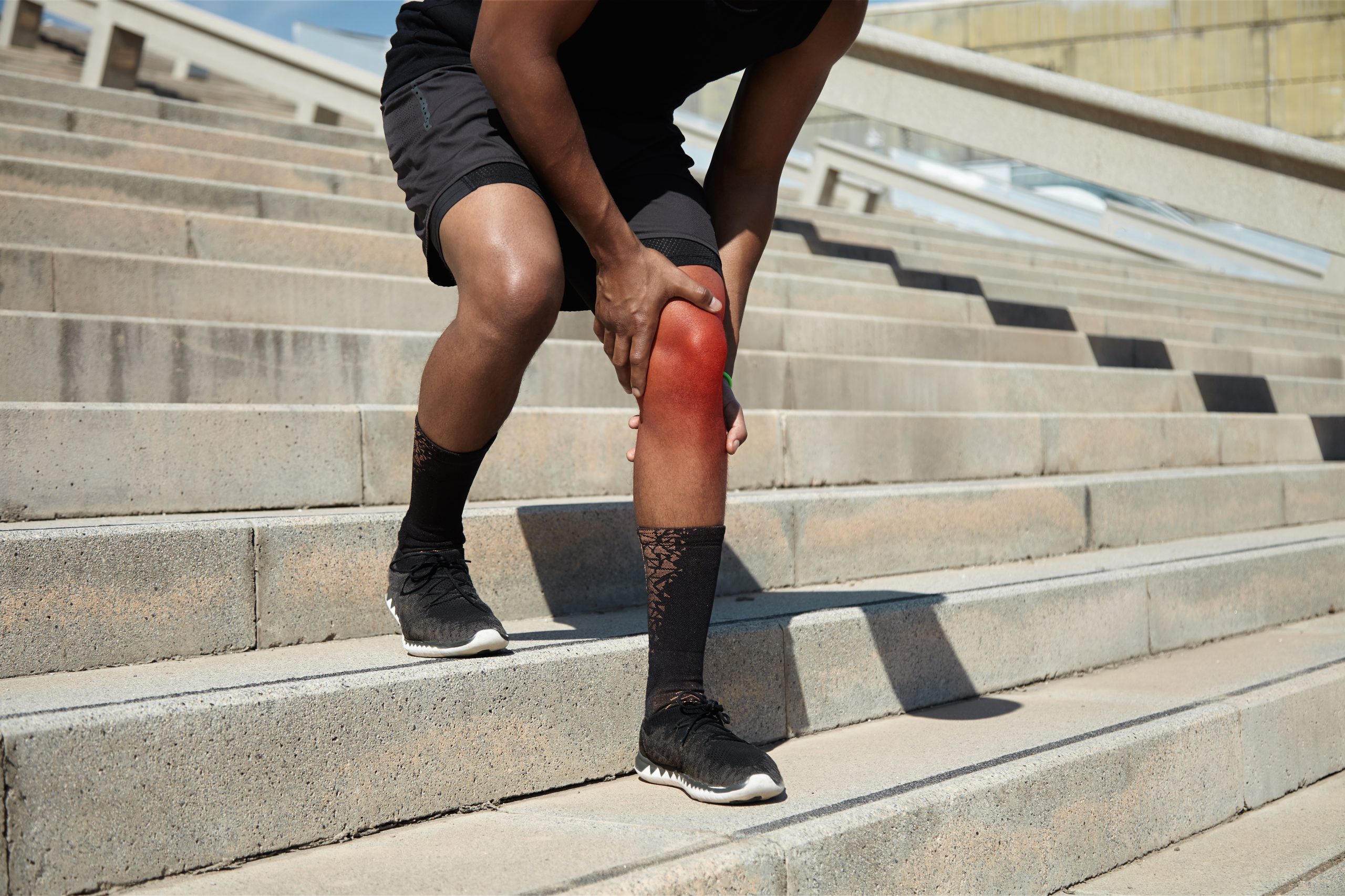 Sports and physical injury concept. Cropped shot of African jogger in black sportswear and running shoes having twitch or sprain in his red knee, standing outdoors, rubbing sore area with his hands